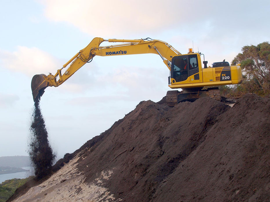 A photo of a yellow excavator placing topsoil containing seed over the disturbed ground.
